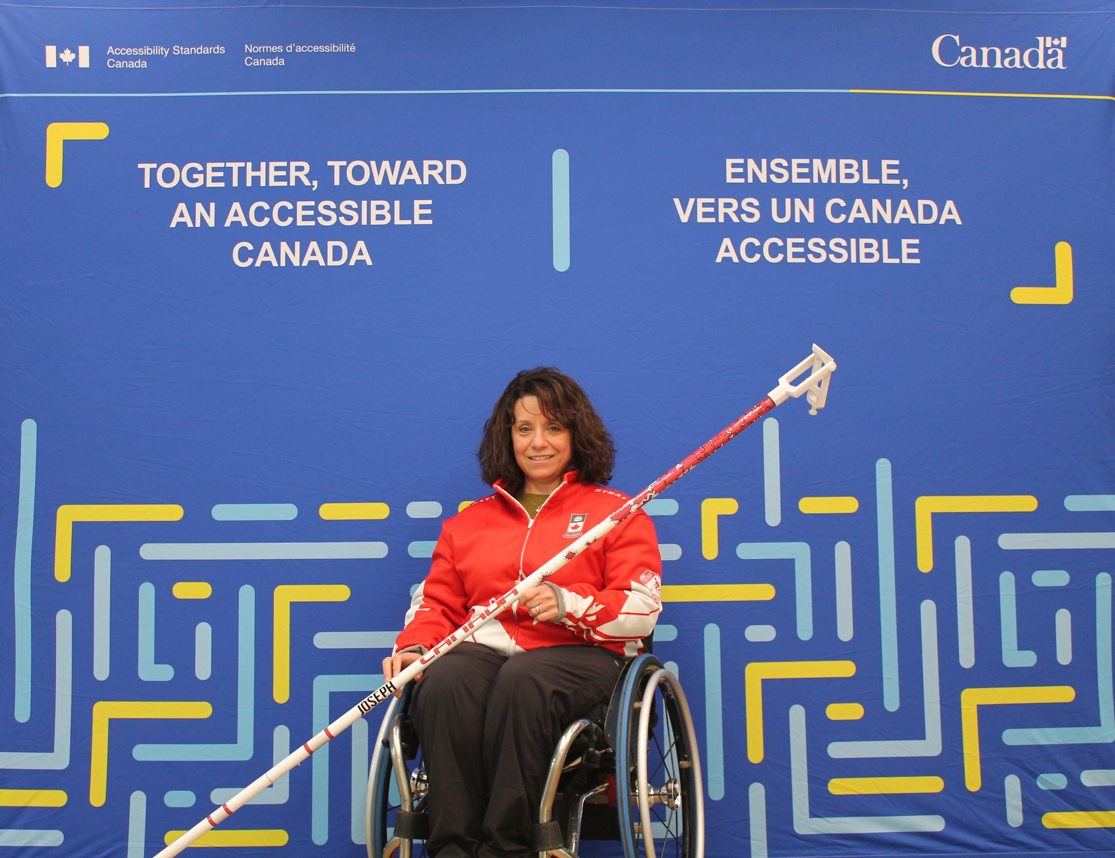 Collinda Joseph poses with her wheelchair and her curling stick. The stick has an adjustable handle at the end that grips the stone and allows her to throw it from a distance. She is in front of a blue backdrop with Accessibility Standards Canada's logo. The English slogan on it reads "Together, toward an accessible Canada". The French slogan on it reads "Ensemble vers un Canada Accessible".