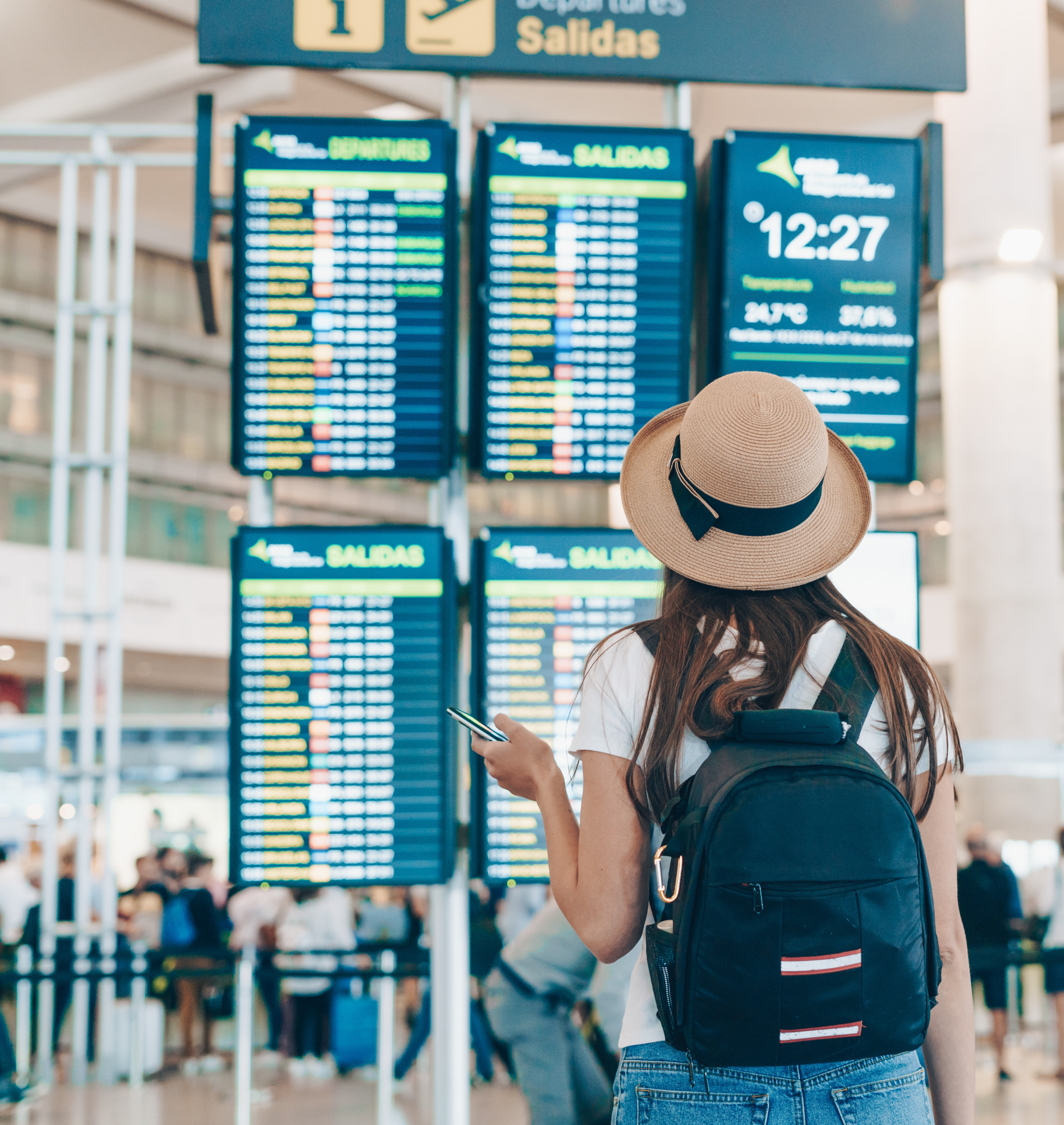 A woman wearing a brown hat and a backpack stands in front of the departure screen in an airport.