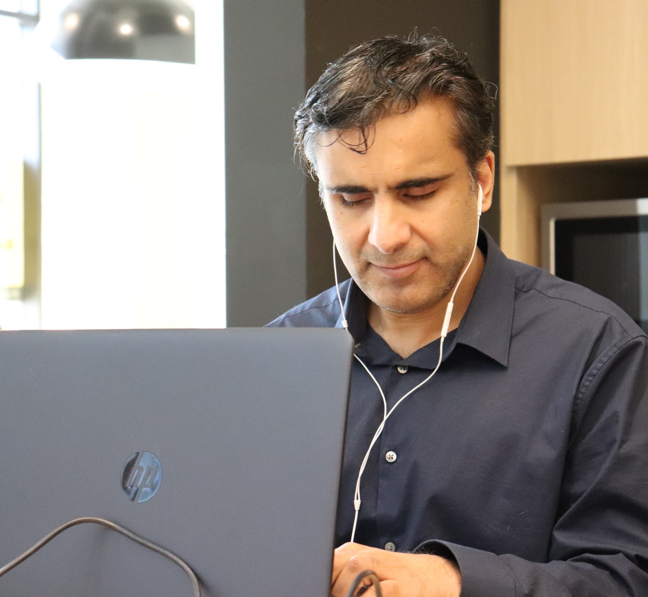 A man with a navy blue shirt is typing on a computer. He has earphones in his ears.
