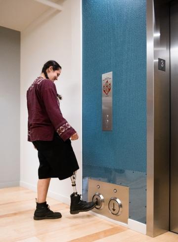An Indigenous Two-Spirit person calls the elevator up by tapping the floor button with their prosthetic leg. Image credit: Attributed to Disabled And Here.